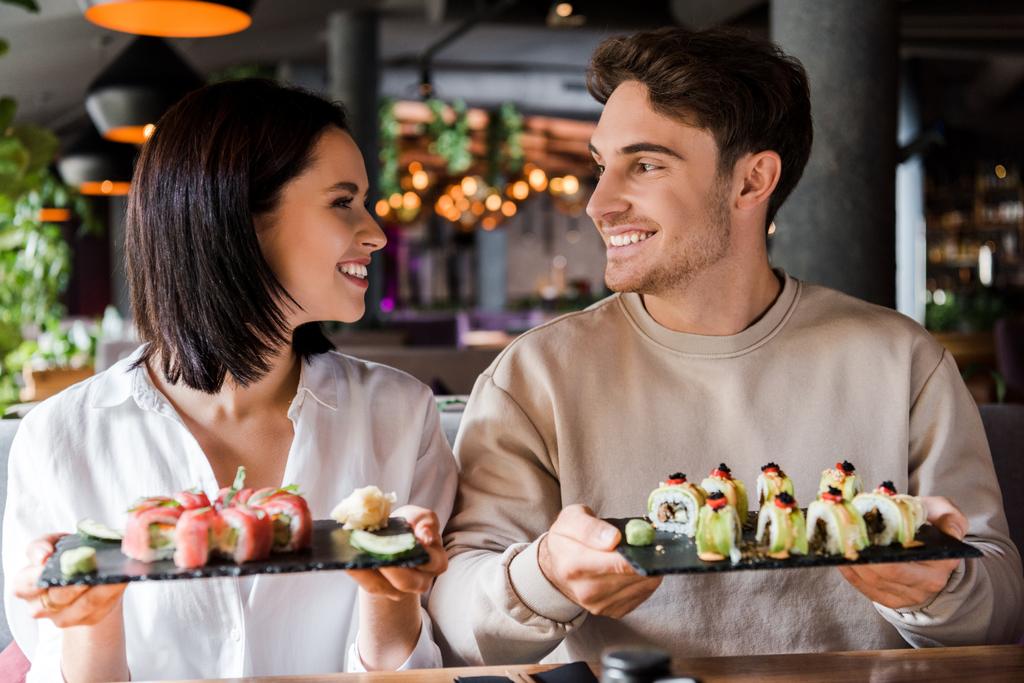 glücklicher Mann und fröhliche Frau lächeln, während sie Teller mit Sushi in der Hand halten  - Foto, Bild