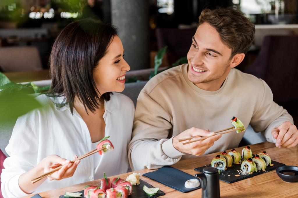 ευτυχισμένος άνθρωπος και χαρούμενη γυναίκα κρατώντας ξυλάκια με νόστιμο σούσι στο εστιατόριο  - Φωτογραφία, εικόνα
