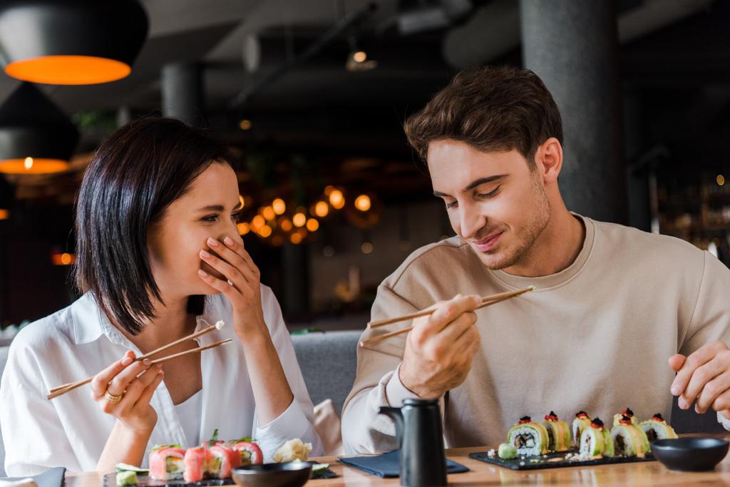 gelukkig man holding eetstokjes in de buurt vrolijk vrouw lachen terwijl cover gezicht in restaurant  - Foto, afbeelding