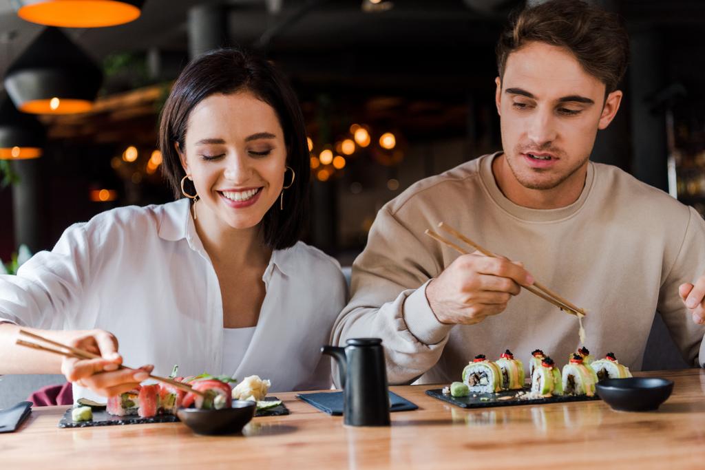 enfoque selectivo de hombre joven sosteniendo palillos cerca de la mujer feliz en el bar de sushi
 - Foto, Imagen