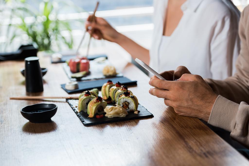 przycięte widok mężczyzna gospodarstwa smartfon w pobliżu młoda kobieta z pałeczki w sushi bar - Zdjęcie, obraz