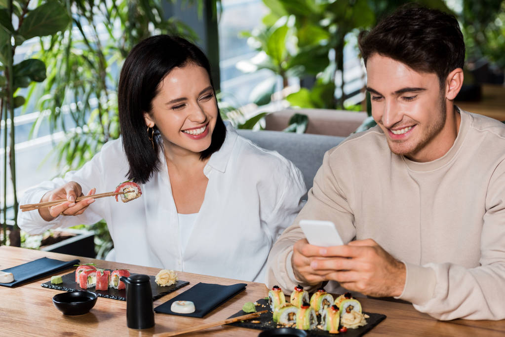 glückliche Frau hält Essstäbchen mit Sushi in der Hand und schaut fröhlichen Mann mit Smartphone an  - Foto, Bild