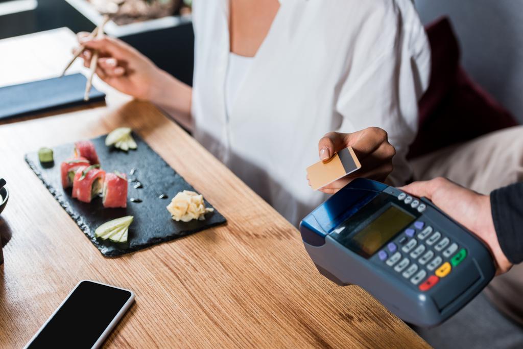 クレジットカードで支払う女性の近くにクレジットカードリーダーを保持するウェイターのクロップドビュー  - 写真・画像