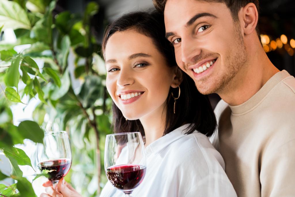 selectieve focus van knappe man met glas met rode wijn in de buurt van positieve vriendin - Foto, afbeelding