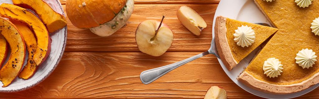 panoramiczny strzał pysznego ciasta dyniowego z bitą śmietaną w pobliżu surowych i pieczonych dyń, i pokrojone jabłko na pomarańczowym drewnianym stole - Zdjęcie, obraz