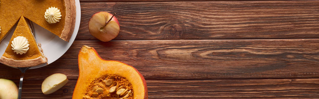 panoramatický záběr lahodného dýňového koláče se šlehačkou v blízkosti řezu a celých jablek, půlka syrové dýně na hnědém dřevěném stole - Fotografie, Obrázek
