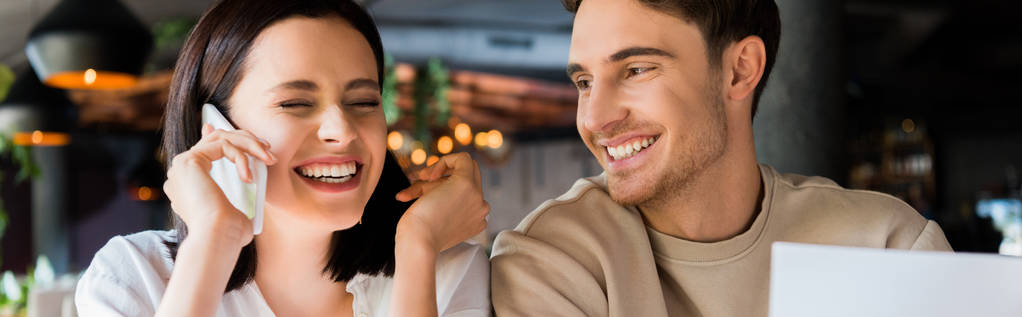 panoramiczne ujęcie szczęśliwego mężczyzny w pobliżu wesołej kobiety rozmawiającej na smartfonie  - Zdjęcie, obraz