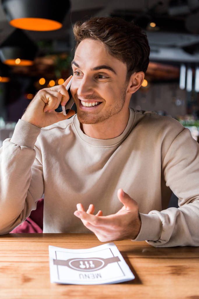 happy man gesturing while talking on smartphone near menu on table  - Photo, Image