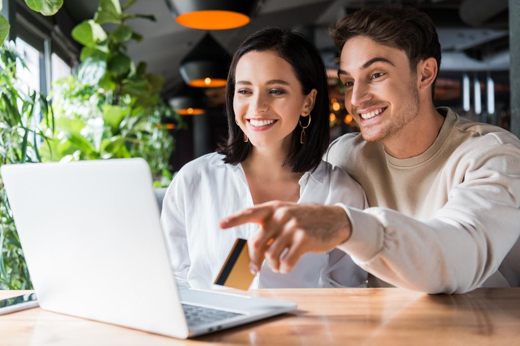 selective focus of happy woman looking at laptop man holding credit card and pointing with finger  - Photo, Image