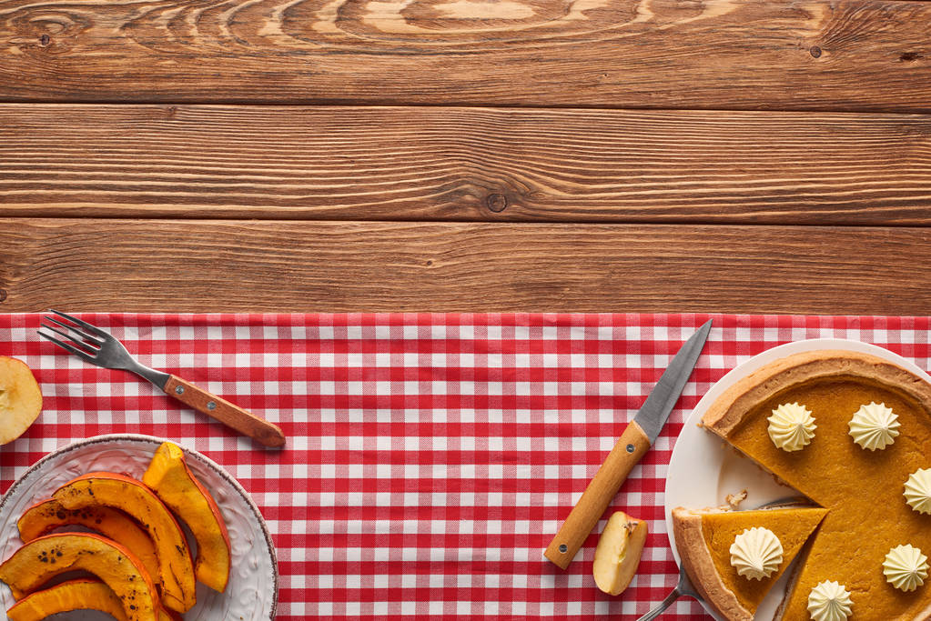 cut pumpkin pie near plate with sliced baked pumpkin, fork and knife on plaid tablecloth on wooden table - Photo, Image