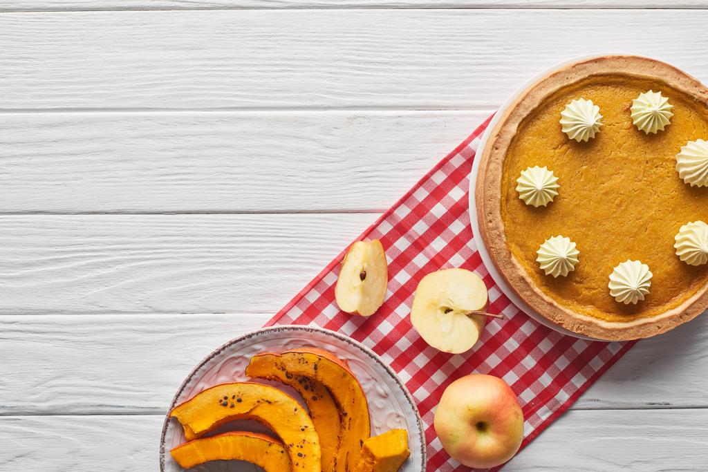 köstlicher Kürbiskuchen mit Schlagsahne auf karierter Serviette neben gebackenem Kürbis, geschnitten und ganzen Äpfeln auf weißem Holztisch - Foto, Bild