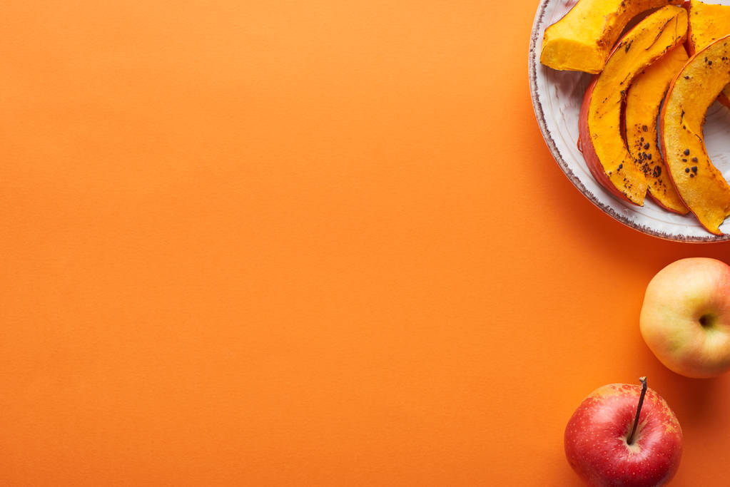In Scheiben geschnittener gebackener Kürbis neben ganzen Äpfeln auf orangefarbener Oberfläche mit Kopierfläche - Foto, Bild