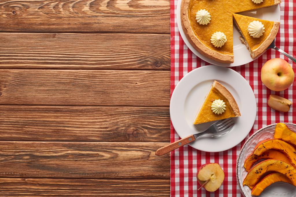delicious pumpkin pie with whipped cream on checkered tablecloth near baked pumpkin, fork, cut and whole apples on brown wooden table - Photo, Image