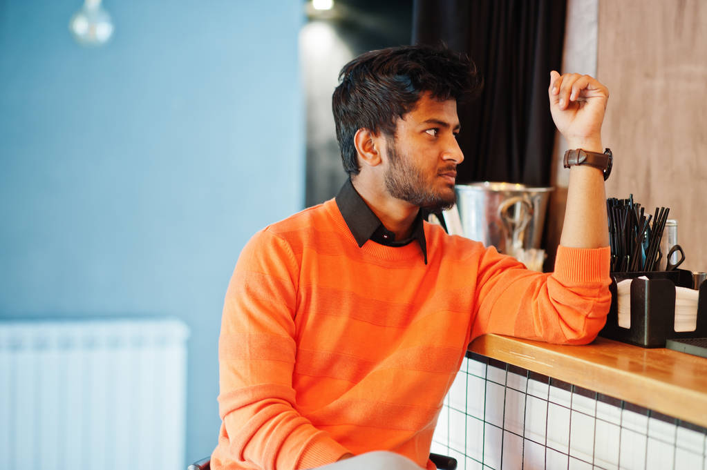 Σίγουροι νέος Ινδός άντρας σε πορτοκαλί πουλόβερ καθιστό πάγκο - Φωτογραφία, εικόνα