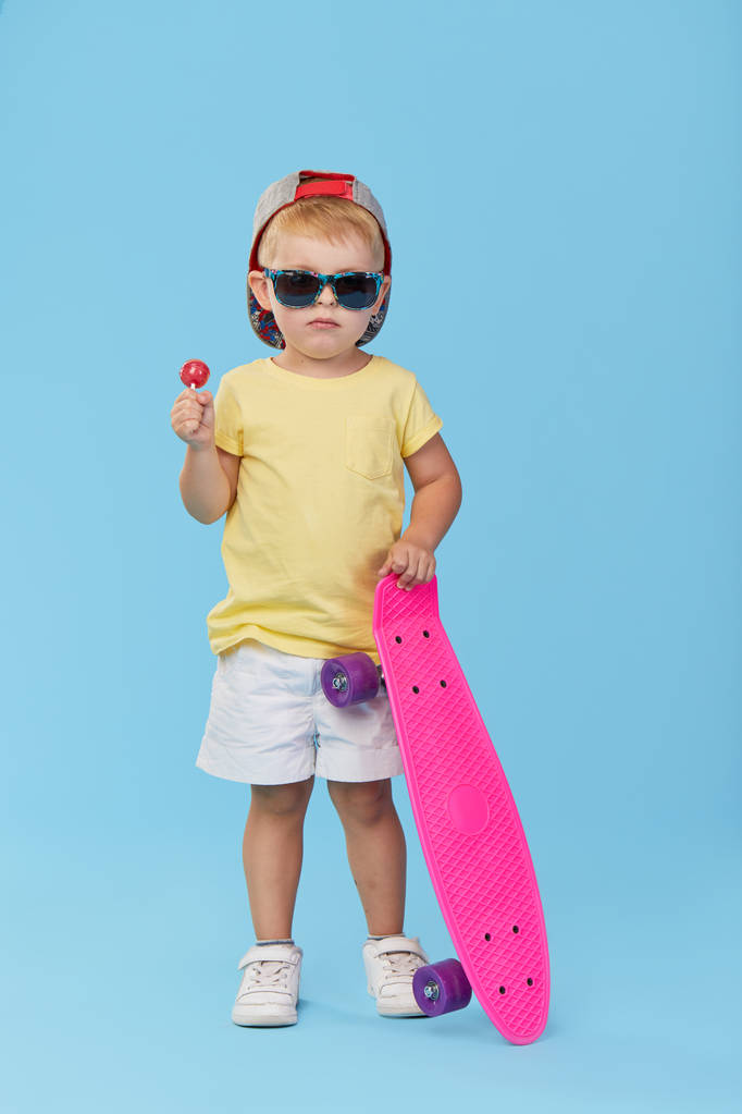 stylisches kleines Kleinkind in Brille mit Skateboard, das sich vor blauem Hintergrund amüsiert - Foto, Bild