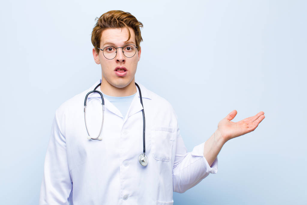 jeune médecin en chef rouge regardant surpris et choqué, avec la mâchoire tombée tenant un objet avec une main ouverte sur le côté contre le mur bleu
 - Photo, image