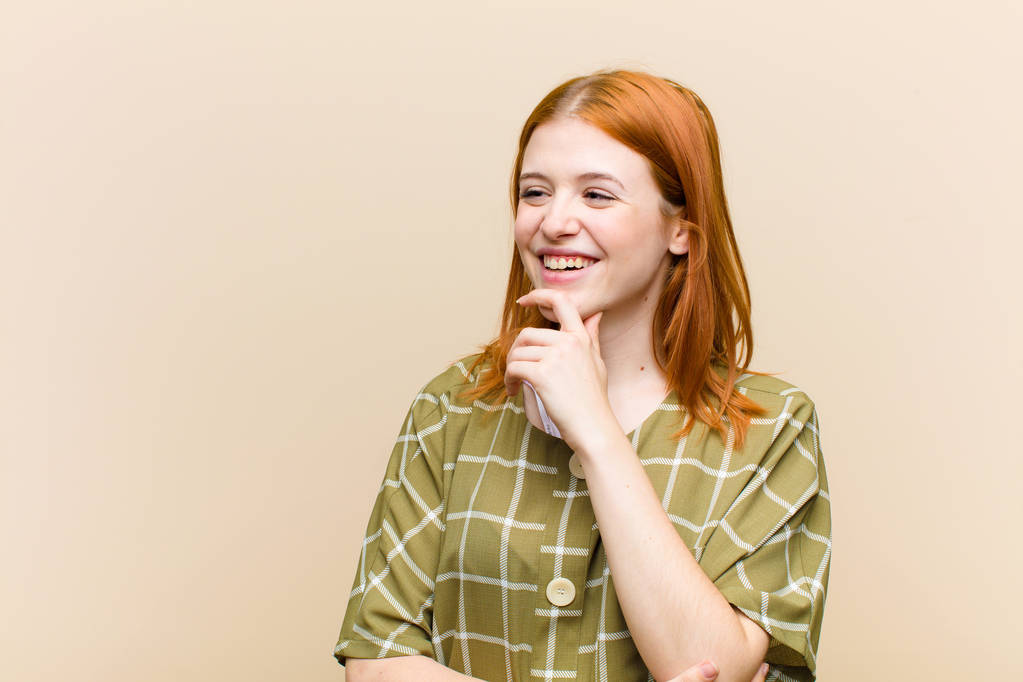 νεαρή κοκκινομάλλα όμορφη γυναίκα χαμογελά με μια ευτυχισμένη, σίγουρη έκφραση με το χέρι στο πηγούνι, αναρωτιούνται και κοιτάζοντας στο πλάι - Φωτογραφία, εικόνα