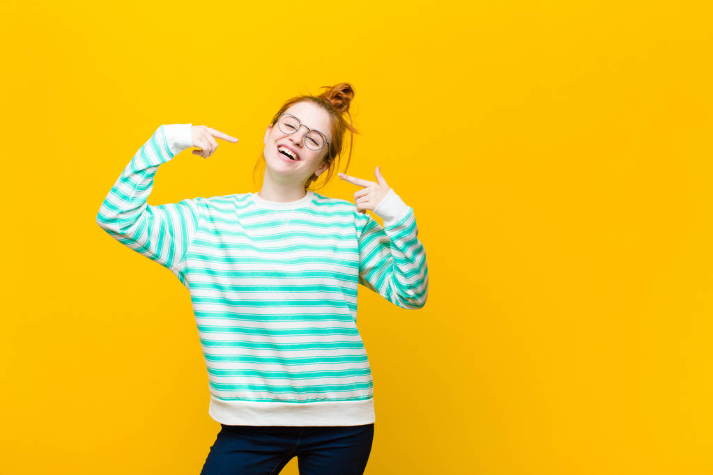 joven mujer pelirroja sonriendo con confianza señalando a su propia sonrisa amplia, actitud positiva, relajada y satisfecha contra la pared naranja
 - Foto, imagen
