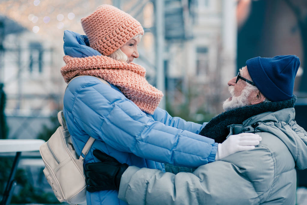 Açık havada duran kışlık kıyafetler giyen güzel birkaç emekli. Ellerini kocasının omuzlarında geçiren kadın - Fotoğraf, Görsel