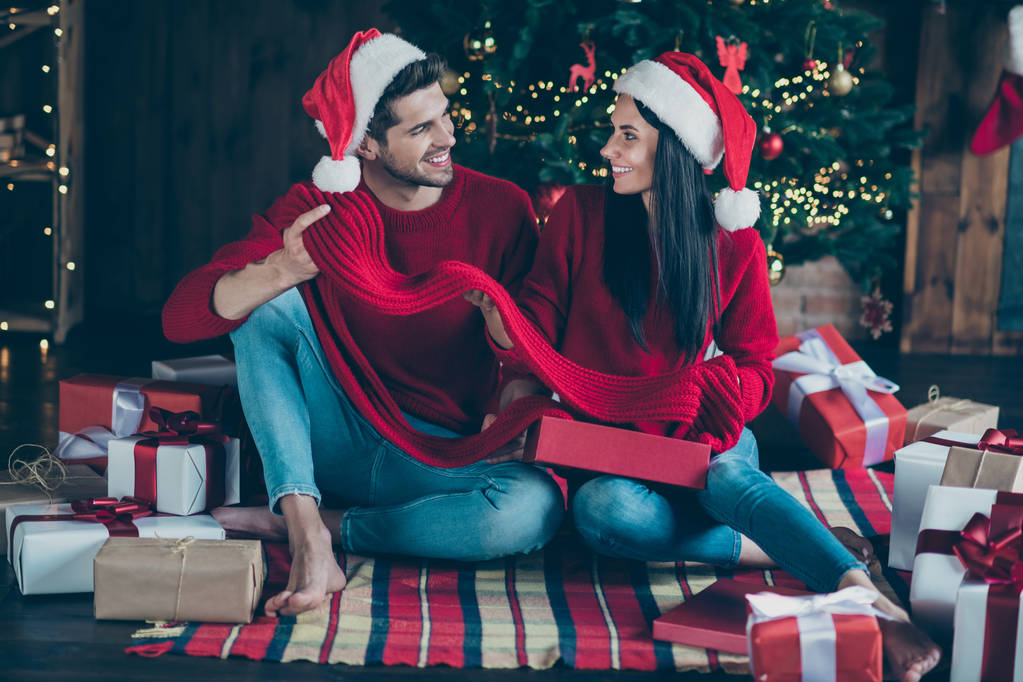 帽子をかぶった2人の陽気な恋人のフルボディ写真は、屋内でX-masの装飾ライトイルミネーションと家の中でクリスマスプレゼントを楽しむチェック柄の毛布の上に座ります - 写真・画像