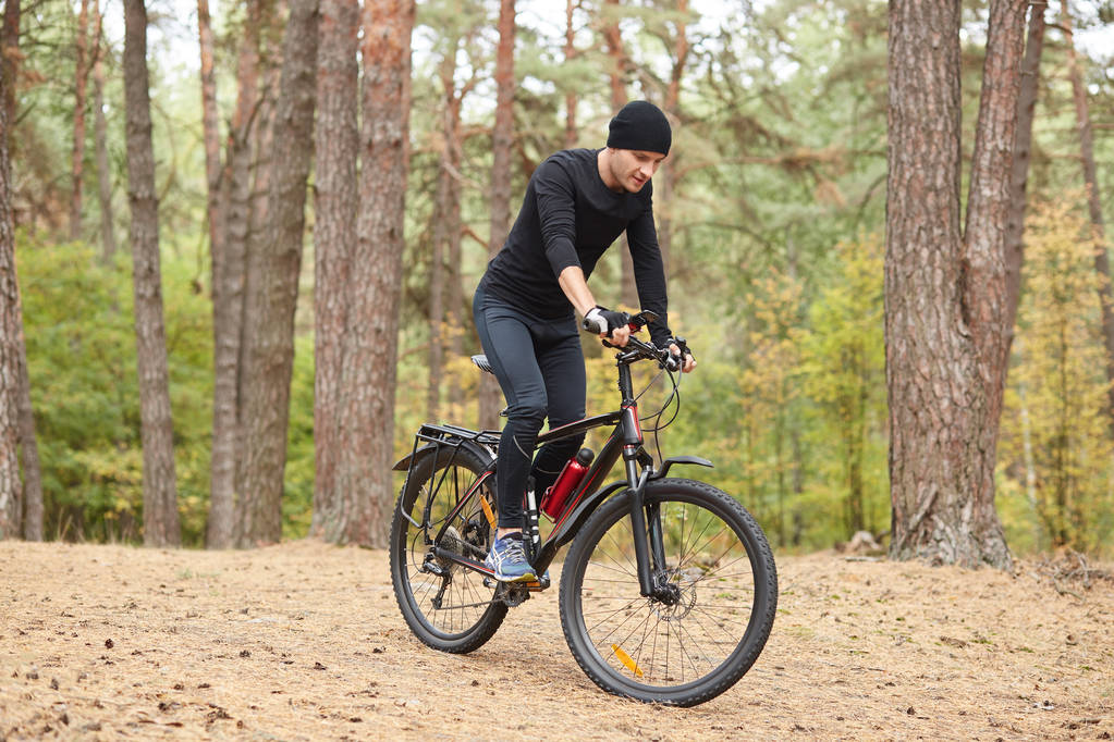 Photo of cyclist on forest trail, trip on bikes in lush green nature, young attractive guy wearing black sportwear rides around trees in forest, spends free time in open air. Healthy lifestyle concept - Photo, Image