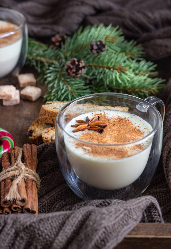 Du lait de poule. Cocktail de lait de Noël à la cannelle, servi dans deux tasses en verre sur un plateau en bois avec biscottes, épices d'hiver, bonbons et branche de sapin
. - Photo, image