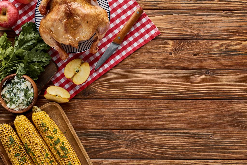 ylhäältä näkymä paahdettua kalkkunaa, grillattua maissia, omenoita, persiljaa ja keltaisia villikukkia punaisella ruudullisella lautasliinalla puupöydällä
 - Valokuva, kuva