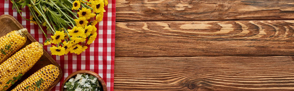 κορυφαία άποψη του ψητού καλαμποκιού και κίτρινα αγριολούλουδα σε κόκκινο καρό χαρτοπετσέτα σε ξύλινο τραπέζι, πανοραμική λήψη - Φωτογραφία, εικόνα