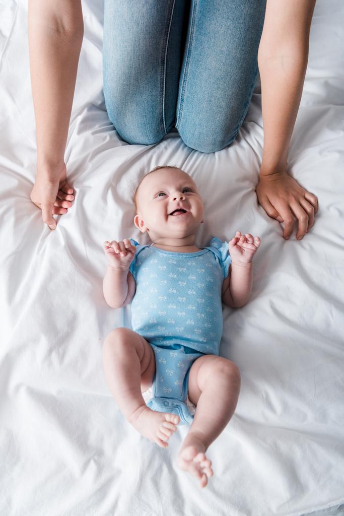 青いベビーボディスーツを着た幸せな赤ちゃんの近くに座っているジーンズの母親の  - 写真・画像