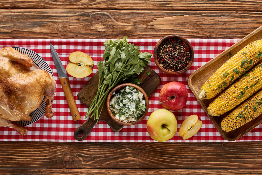 τοπ άποψη της ψητής γαλοπούλας, ψητό καλαμπόκι και μήλα με μαϊντανό και πιπέρι που σερβίρεται σε ξύλινο τραπέζι με κόκκινη πετσέτα - Φωτογραφία, εικόνα