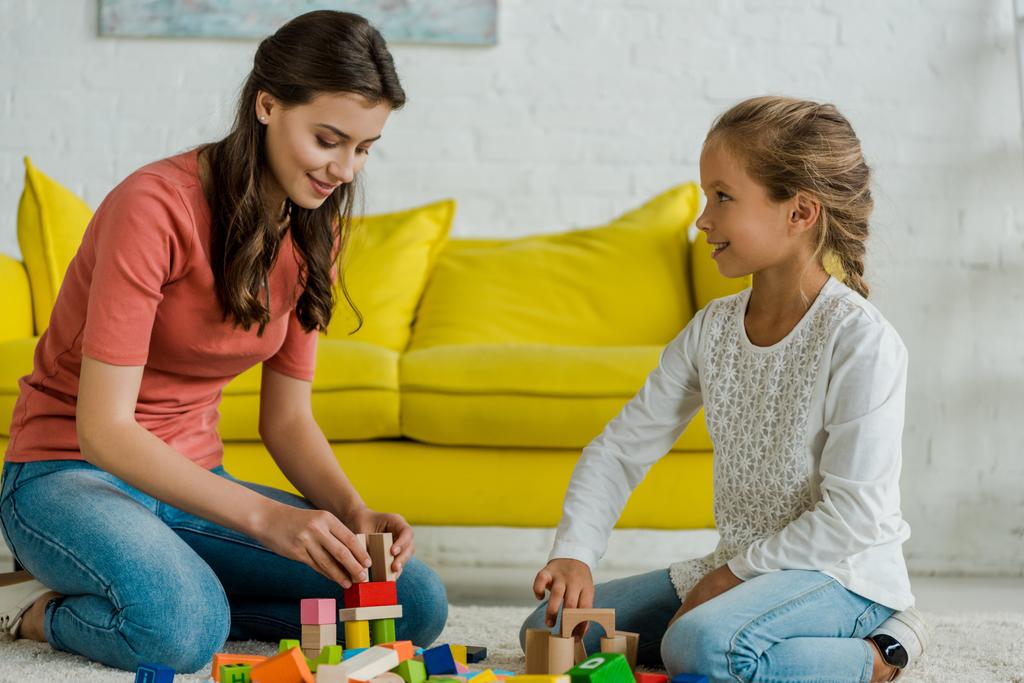 glückliches Kind schaut Babysitter in der Nähe von Spielzeugklötzen auf Teppich an  - Foto, Bild