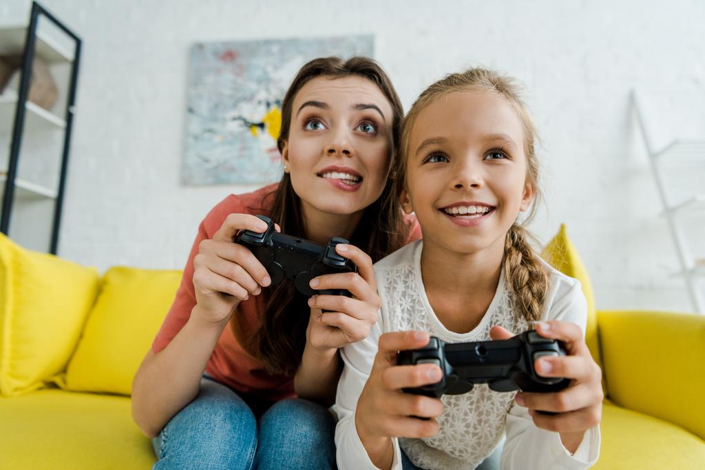KYIV, UKRAINE - SEPTEMBER 4, 2019: babysitter biting lips near happy kid while playing video game in living room  - Photo, Image