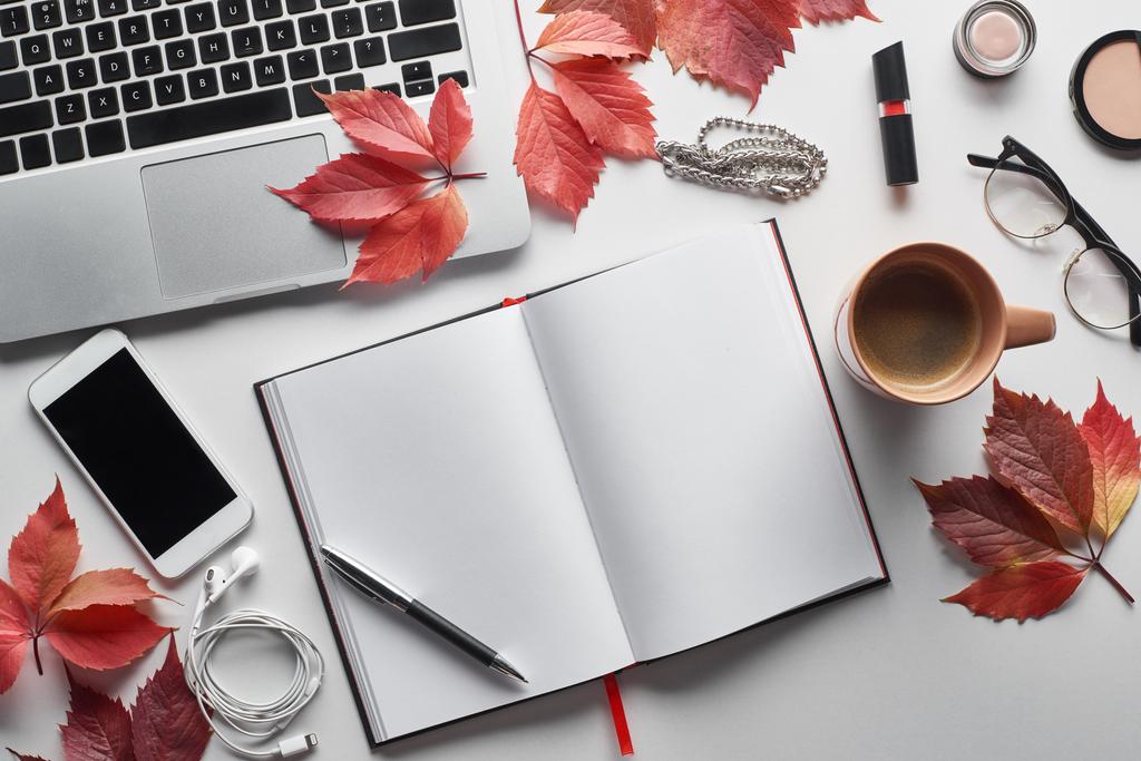 bovenaanzicht laptop bij smartphone, koffiebeker, cosmetica, oortjes, glazen, notebook en rode bladeren van wilde druiven op witte tafel - Foto, afbeelding
