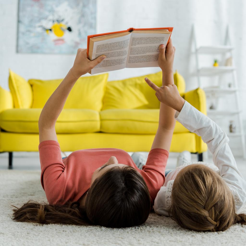 niño señalando con el dedo en el libro mientras está acostado en la alfombra con la niñera
  - Foto, Imagen