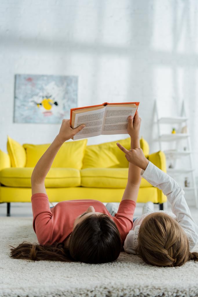 child pointing with finger at book while lying on carpet with babysitter  - Photo, Image