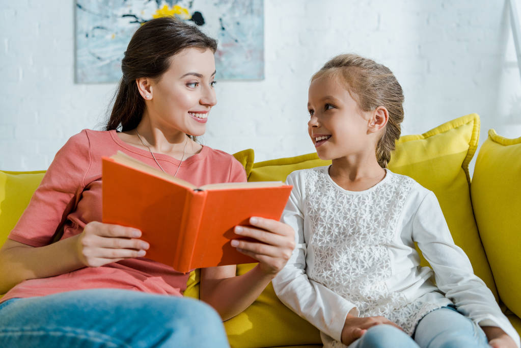 幸せな子供の選択的焦点はソファに座って本を持っているベビーシッターを見て  - 写真・画像
