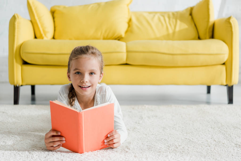 feliz niño sosteniendo libro blanco acostado en la alfombra cerca de sofá amarillo
  - Foto, Imagen