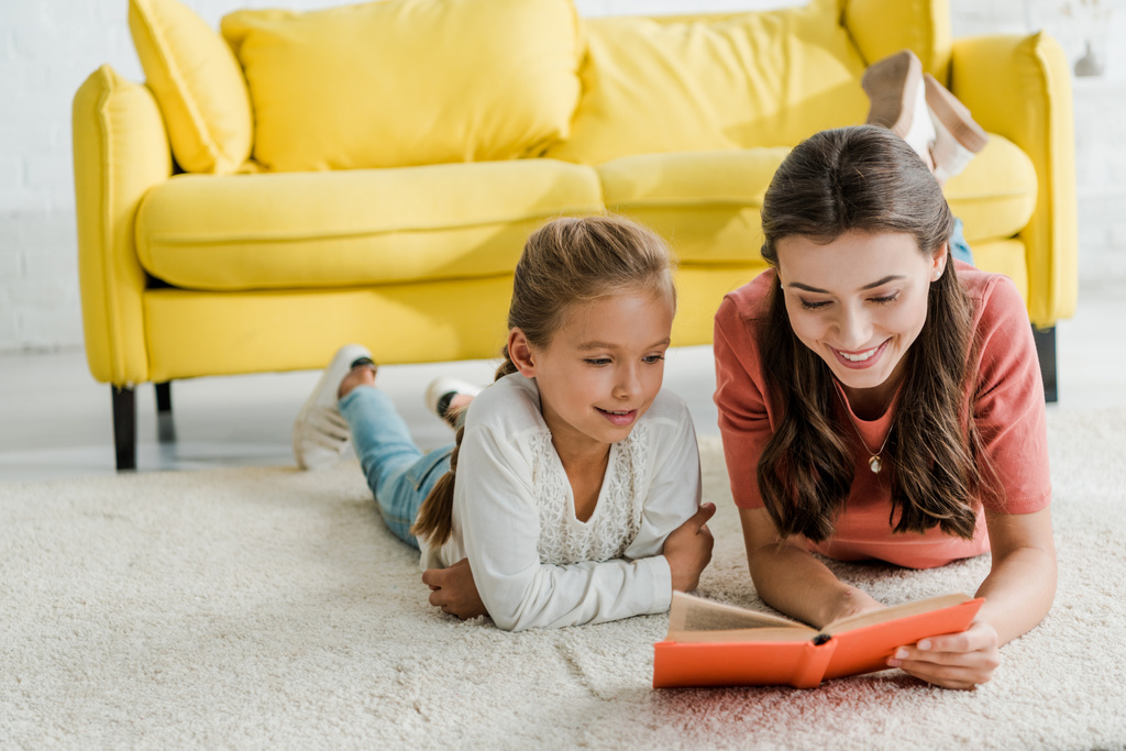 幸せなベビーシッターは本を読んでいる間に笑顔の子供とカーペットの上に横たわる  - 写真・画像