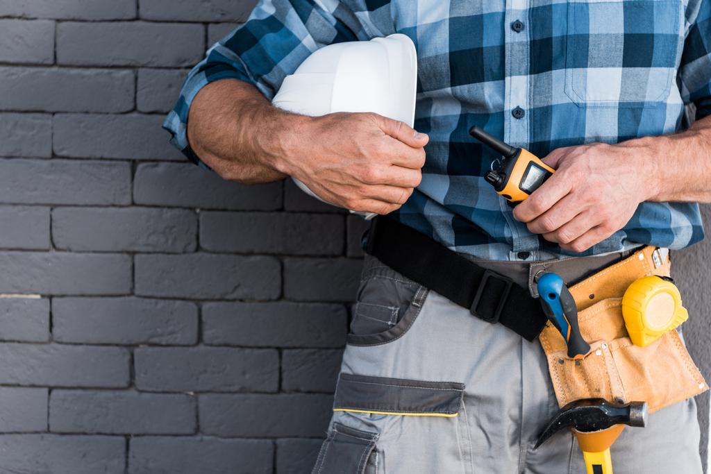 cropped view of builder holding walkie talkie and helmet near brick wall  - Photo, Image