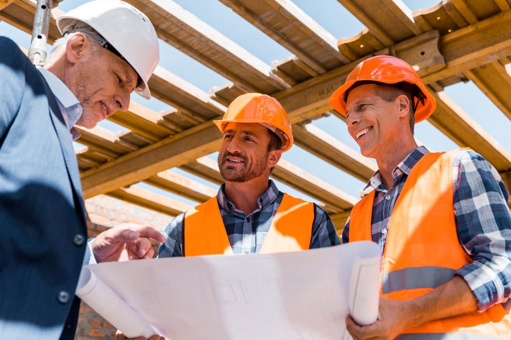 низкий угол обзора строителей рядом со зрелым бизнесменом в шлеме с чертежом
  - Фото, изображение