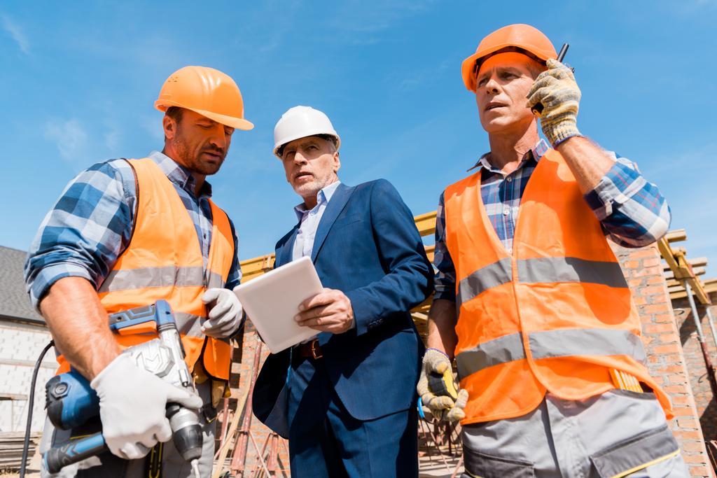 Niedrigwinkel-Ansicht des Bauunternehmers mit Walkie Talkie in der Nähe von Mitarbeiter und Geschäftsmann mit digitalem Tablet  - Foto, Bild