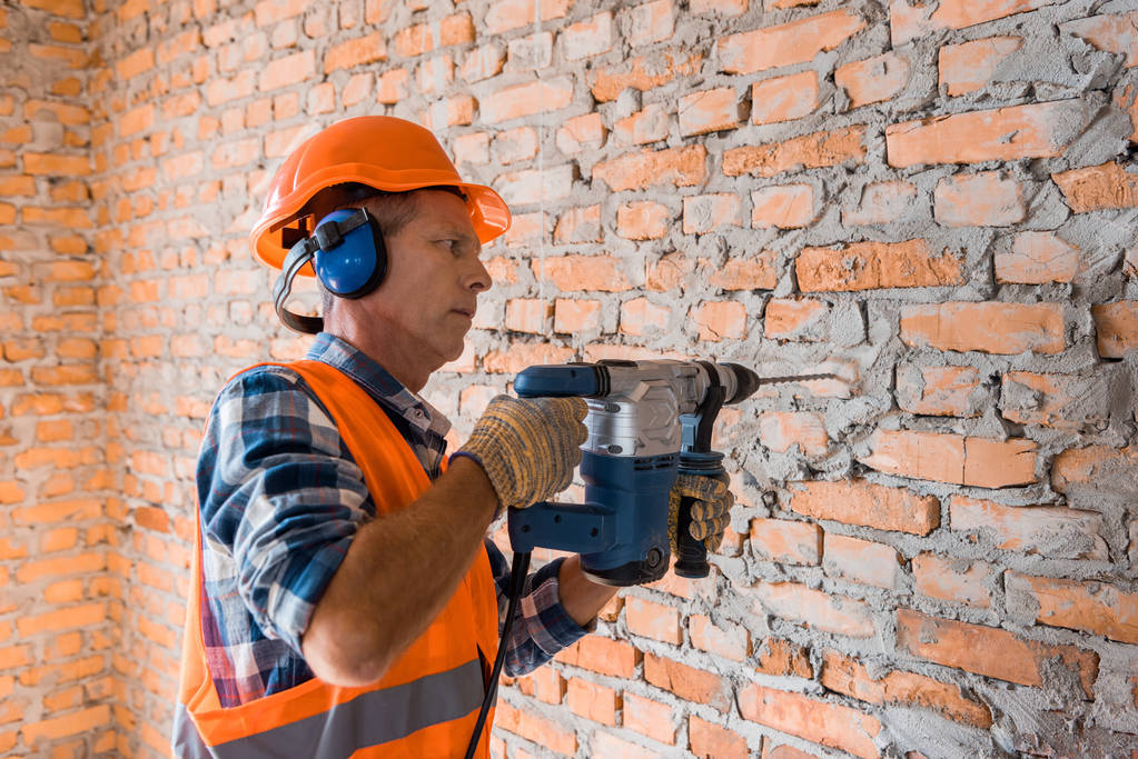 construtor maduro no capacete usando martelo broca perto da parede de tijolo
  - Foto, Imagem