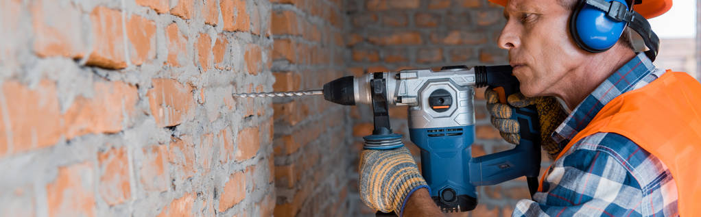 tiro panorâmico de construtor maduro usando broca de martelo perto da parede de tijolo
  - Foto, Imagem