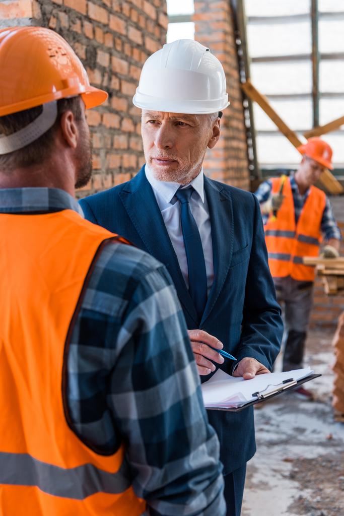селективная направленность бородатого бизнесмена в шлеме, держащего планшет рядом со строителями
  - Фото, изображение