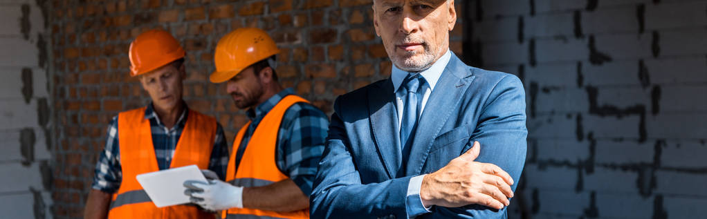 панорамный снимок бизнесмена, стоящего со скрещенными руками рядом со строителями
 - Фото, изображение
