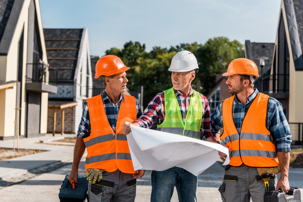 mature builder holding blueprint near coworkers and houses  - Photo, Image