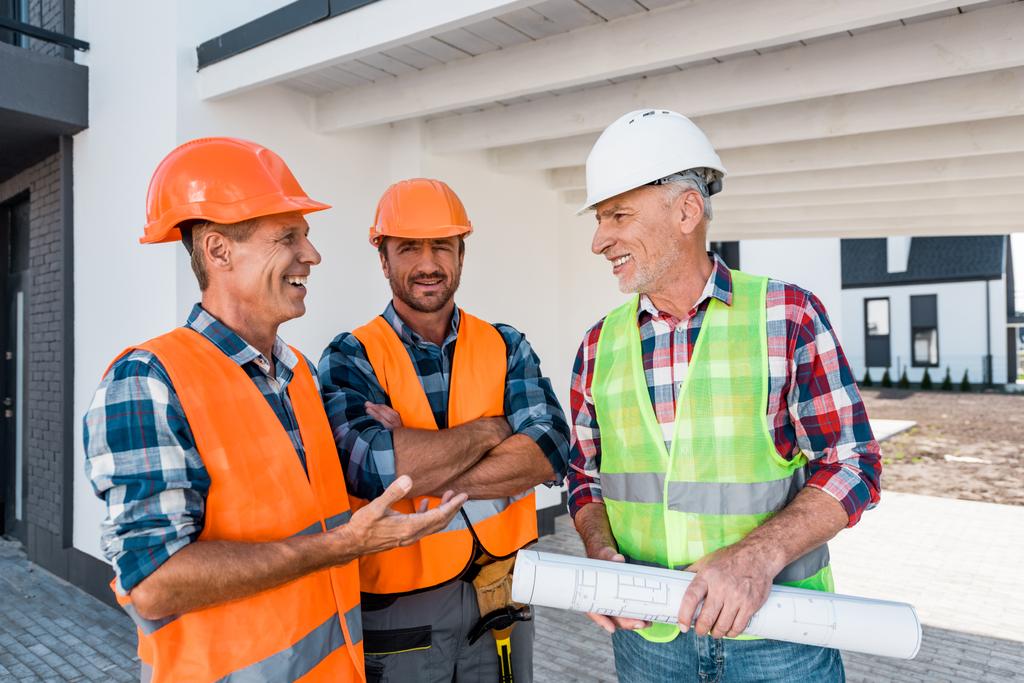 happy constructors in helmets standing near coworker with blueprint  - Photo, Image
