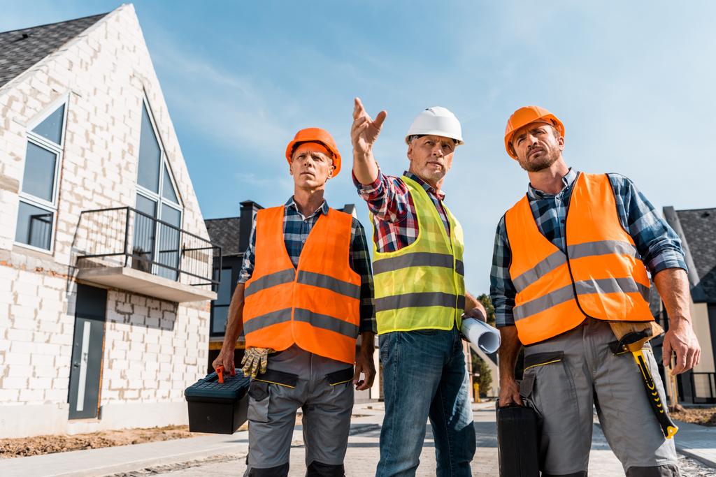 constructors in helmets holding toolboxes near coworker gesturing near houses  - Photo, Image