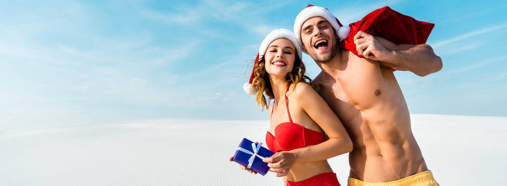 panoramisch schot van sexy en glimlachende vriendin houden geschenk en vriendje met santa zak op het strand in Malediven  - Foto, afbeelding