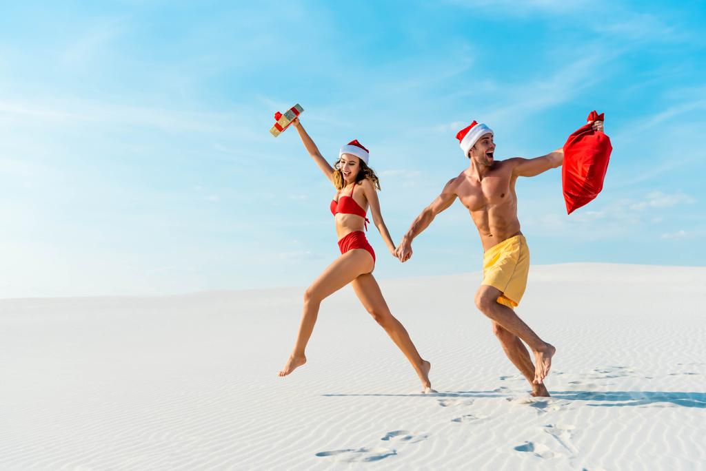 σέξι και χαμογελαστή φιλενάδα κρατώντας δώρο και φίλο με santa σάκο τρέχει στην παραλία στις Μαλδίβες  - Φωτογραφία, εικόνα